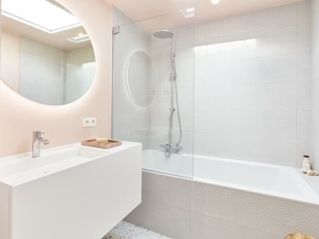 Combinaison bain douche avec baignoire blanche, paroi de bain en verre et colonne de douche chromée à côté d’un meuble de salle de bains blanc avec vasque et miroir rond, dans une salle de bains moderne de x²o salles de bains.