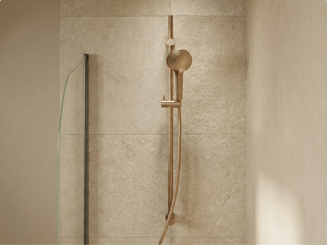 Douchette dorée avec pomme ronde montée sur une barre coulissante verticale contre un mur à texture claire, dans une salle de bains de x²o salles de bains.
