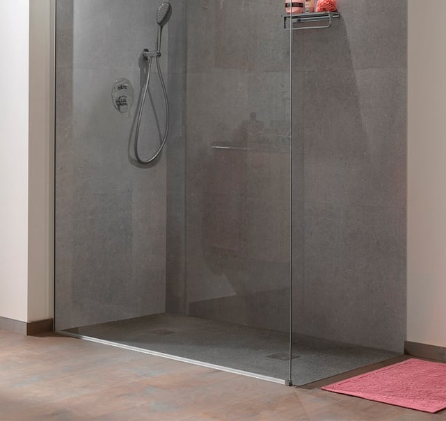 Graue Duschwand mit Glastür, Duschkopf mit Schlauch, Regal mit Produkten an der Wand und rosa Duschmatte auf dem Boden