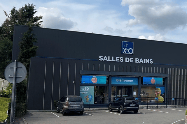 Vue extérieure d’un magasin x²o salles de bains avec façade sombre et grande inscription “SALLES DE BAINS”. Un parking avec plusieurs voitures se trouve devant, et l’entrée affiche “Bienvenue” avec des panneaux promotionnels.
