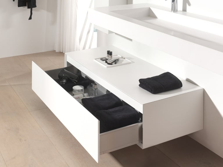 Détail d’un meuble suspendu blanc de X²O salles de bains avec tiroir ouvert contenant serviettes, sèche-cheveux et produits de soin, intégré dans une salle de bain mansardée.