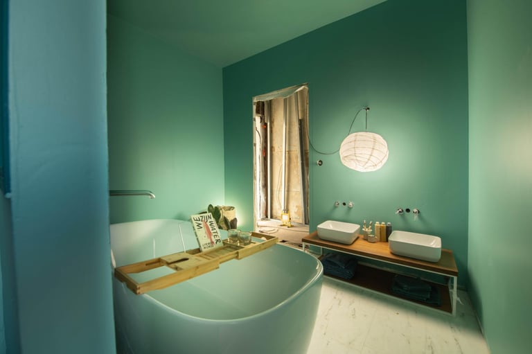 Stilvolles Badezimmer mit grünen Wänden, freistehender Badewanne mit Holzregal, zwei Waschbecken und warmem Licht. Das Fenster ist mit einem Stoff bedeckt