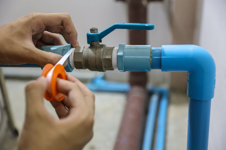 Gros plan d’une installation de plomberie où des mains appliquent du ruban d’étanchéité autour d’un raccord métallique entre des tuyaux bleus. Une vanne d’arrêt est visible, illustrant un travail technique chez x²o salles de bains.