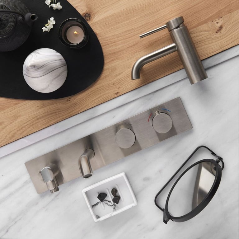 Bovenaanzicht van een inbouwkraan en bedieningsknoppen in geborstelde coolmetal afwerking, gemonteerd in een marmeren wastafelblad met houten accenten. De compositie toont luxe materialen en verfijnde details van x²o badkamers.