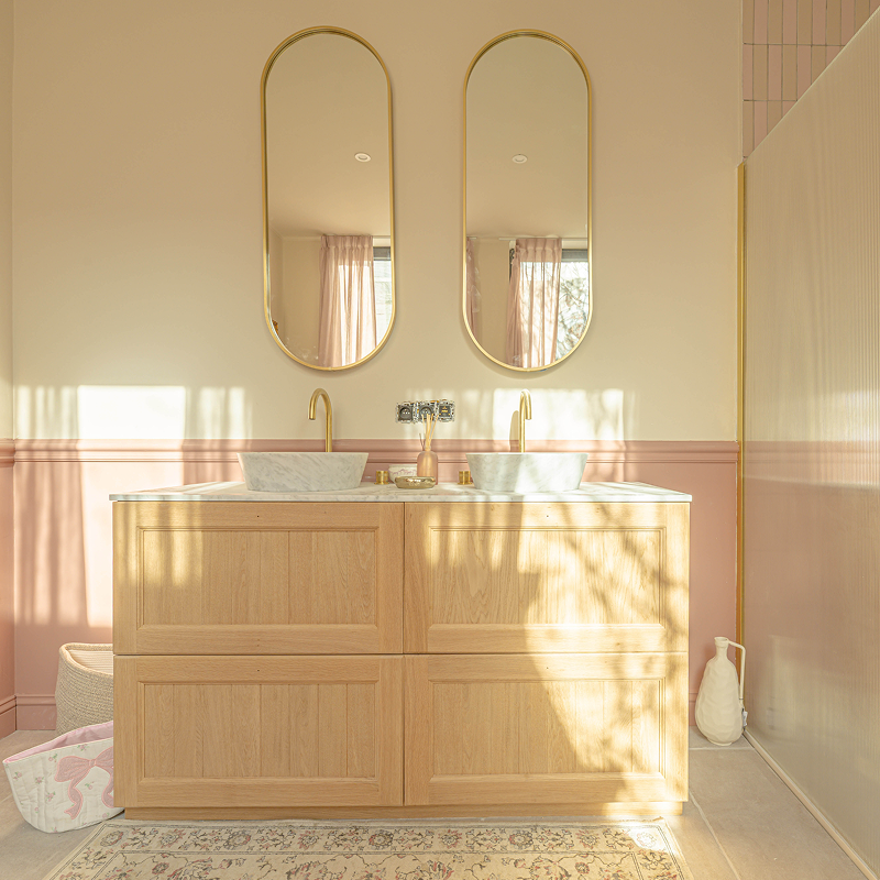 Lichte badkamer met houten dubbel wastafelmeubel, twee hoge ovale spiegels, marmeren waskommen, gouden kranen en roze lambrisering.