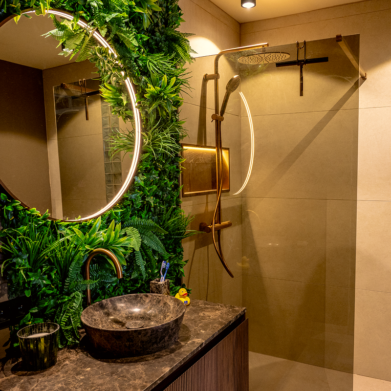 Sfeervolle badkamer met donkere natuurstenen waskom, houten wastafelmeubel, ronde verlichte spiegel, groene plantenwand en inloopdouche met koperkleurige regendouche.