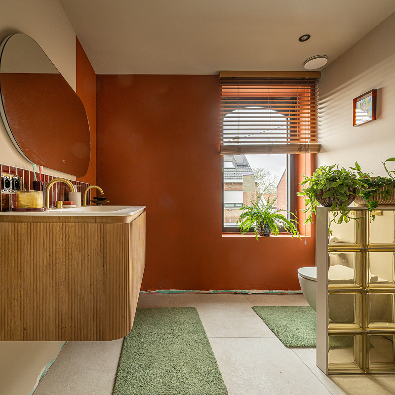Warme badkamer met geribbeld houten wastafelmeubel, organische spiegel, rode accentmuur, houten jaloezieën, planten en glazen bouwstenen.