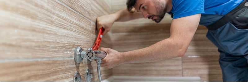 Installateur von x²o Badezimmer, der einen Wasserhahn an einer Wand mit sichtbaren Rohren und Werkzeug montiert.