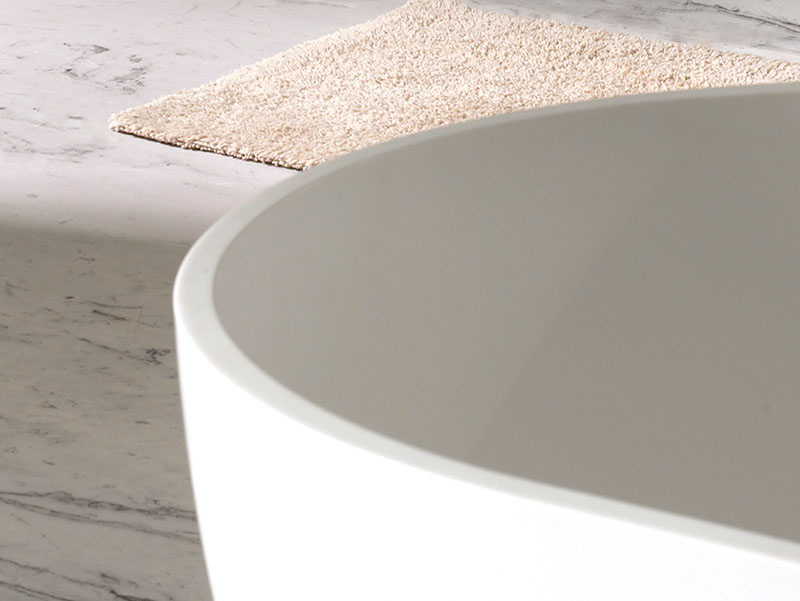 Detail van een vrijstaand bad met afgeronde rand, geplaatst op een marmeren vloer en een beige badmat.