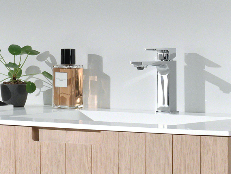 Detail van een houten wastafelmeubel met witte wastafel, chromen kraan, een plant en een parfumfles.