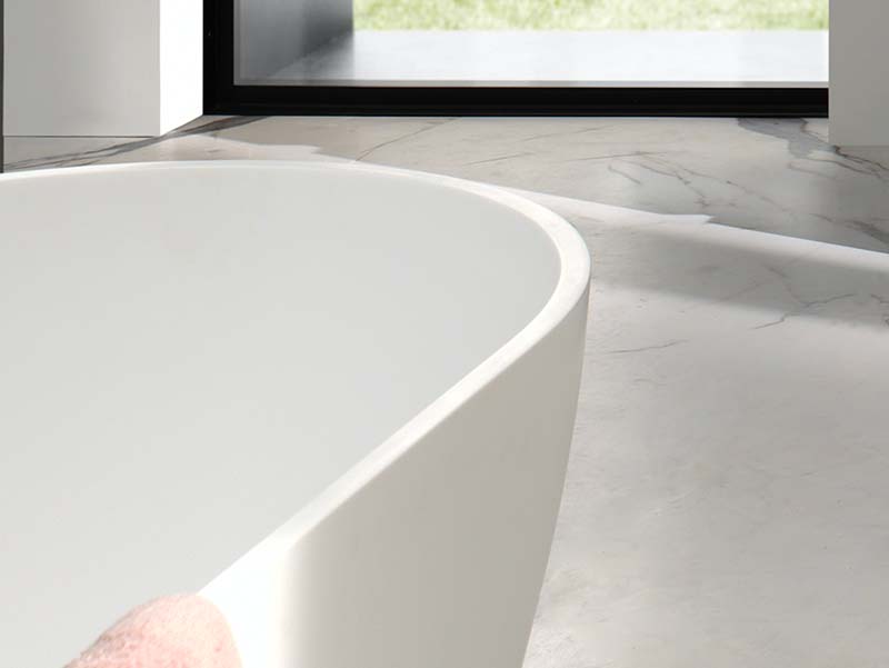 Baignoire îlot blanche aux formes arrondies, posée sur un sol en marbre avec lumière naturelle.