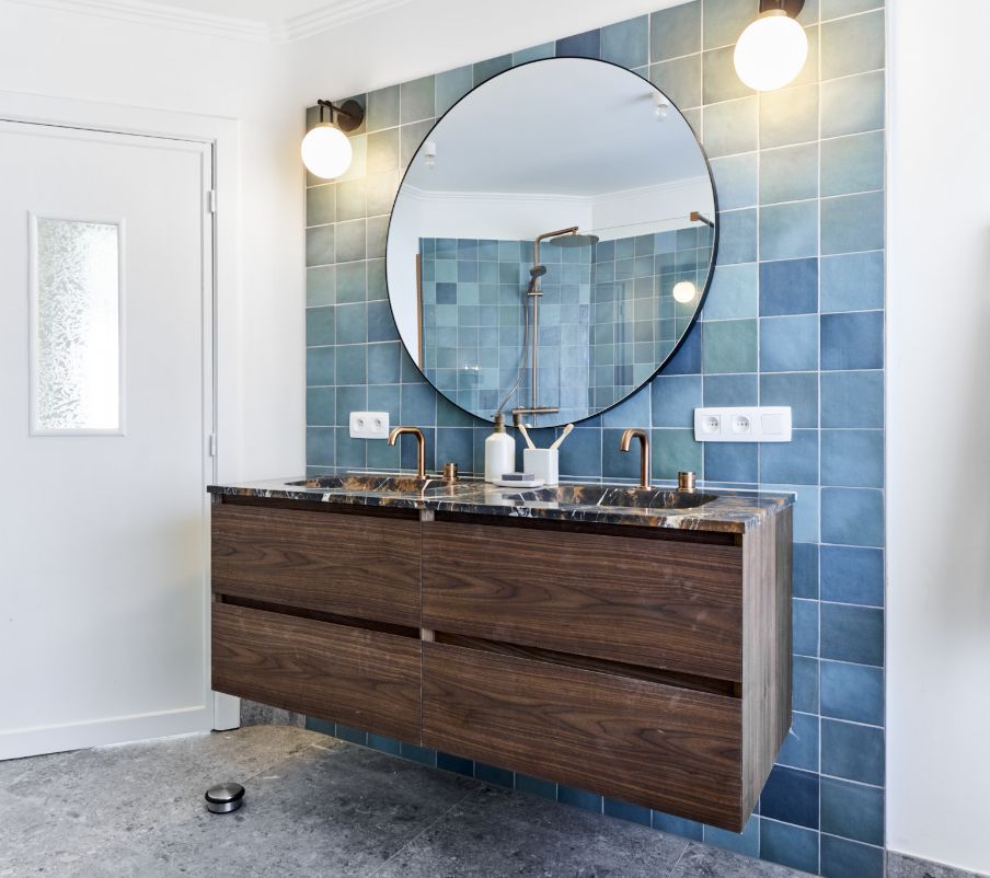 Salle de bains avec un miroir rond, lavabo en marbre, meuble en bois et carrelage bleu sur le mur.