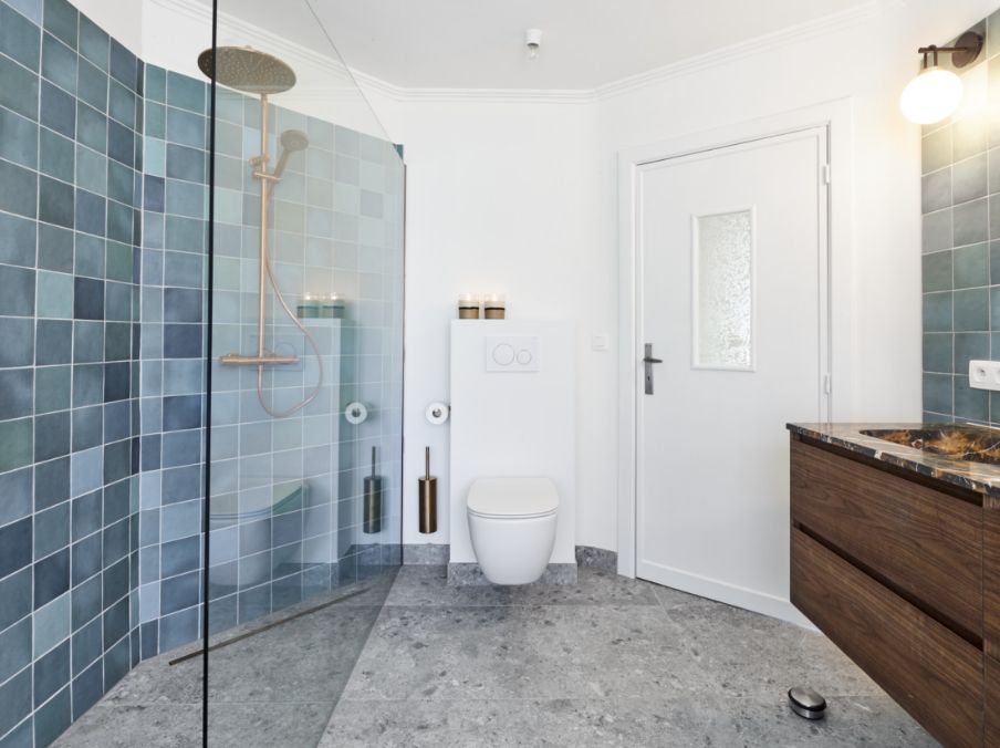 Salle de bains avec une douche en verre, des toilettes, un sol en marbre gris, des carreaux bleus et un meuble lavabo en bois.