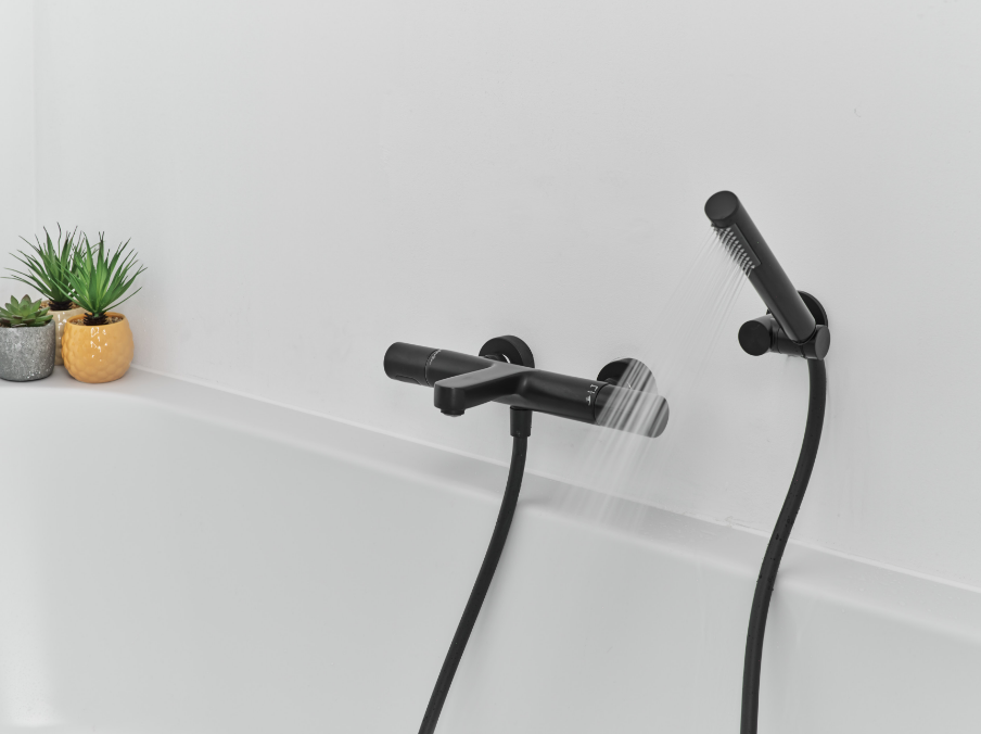 Détail d’un mitigeur de baignoire noir mat de x²o salles de bains avec douchette, installé sur une baignoire blanche, avec un mur minimaliste et de petites plantes décoratives sur le rebord.