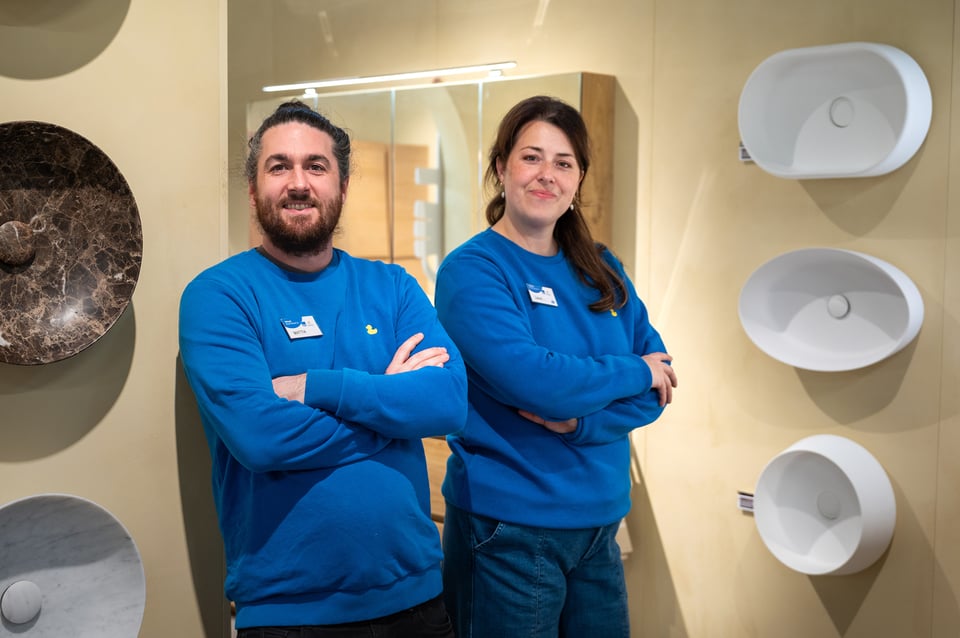Zwei Mitarbeiter von x²o Badezimmer in blauen Pullovern stehen mit verschränkten Armen in einem Badezimmer-Showroom, umgeben von weißen Design-Waschbecken an der Wand und Ausstellungsstücken, mit beleuchtetem Spiegel im Hintergrund