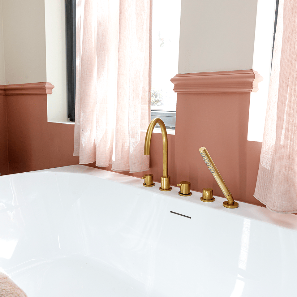 Detail van een vrijstaand wit bad met gouden kraan en handdouche, roze lambrisering en lichte gordijnen.