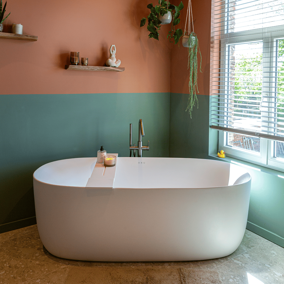 Badkamer met vrijstaand wit bad, terracotta-groene muren, houten jaloezieën, wandplanken, hangplanten en decoratie rond het bad.