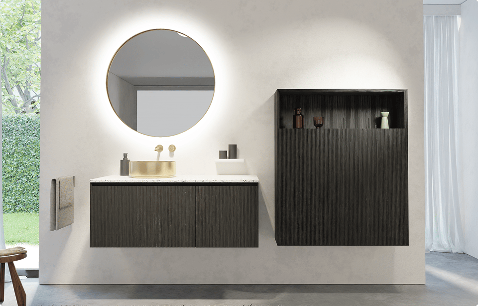 Storke badkamermeubel in donker geribbeld hout met wit terrazzo wastablet, ronde champagnegouden opbouw waskom en bijpassende kraan, geplaatst onder een ronde spiegel met geïntegreerde verlichting en naast een open wandnis met decoratieve accessoires in een badkamer van x²o badkamers.