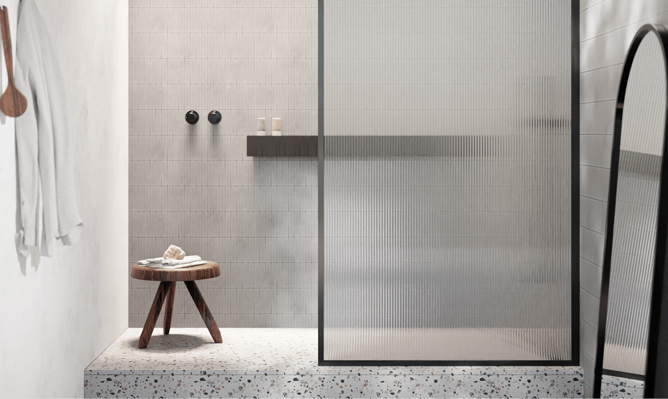 Douche à l’italienne moderne d’une salle de bains x²o, posée sur une plateforme en terrazzo, avec des carreaux muraux gris clair posés à la verticale et une paroi de douche en verre nervuré encadrée de noir. Un petit tabouret en bois avec une serviette est visible à gauche, et un grand miroir debout à droite.