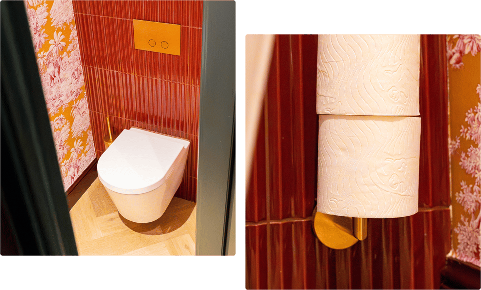 Een wit hangtoilet van x²o badkamers tegen een wand met verticale rode tegels en oranje-roze behang met tropische motieven, gecombineerd met een goudkleurige toiletborstel en een inbouwbedieningspaneel.