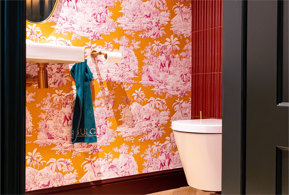 Gouden toiletrolhouder van x²o badkamers met dubbele toiletrollen tegen een rode wandtegel en roze behang.