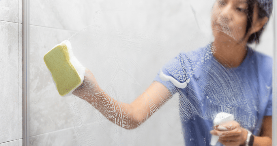 Femme portant un haut bleu qui nettoie une paroi de douche en verre avec une éponge jaune et de la mousse, tenant un produit de nettoyage dans l’autre main, dans une salle de bains de x²o salles de bains.