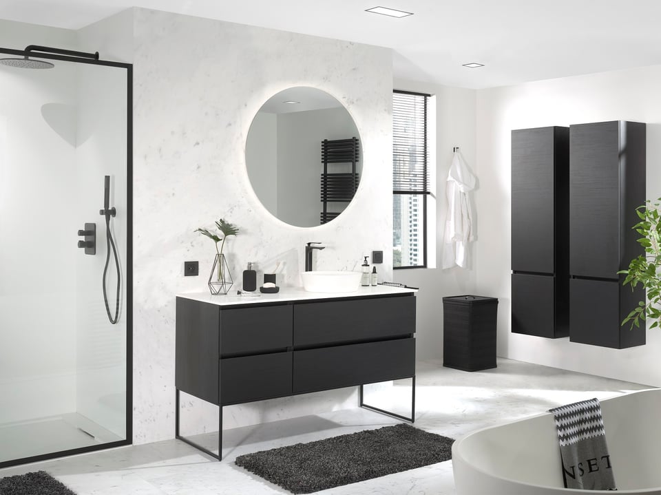 Moderne complete badkamer met een zwarte houten wastafelmeubel op slank metalen frame, een witte opbouwwaskom en een ronde spiegel met indirecte LED-verlichting tegen een marmerlook wand. Links is een inloopdouche met zwart aluminium profiel en regendouche zichtbaar, terwijl rechts hoge zwarte opbergkasten, een raam met jaloezie en een vrijstaand bad zorgen voor een strak en luxueus geheel.
