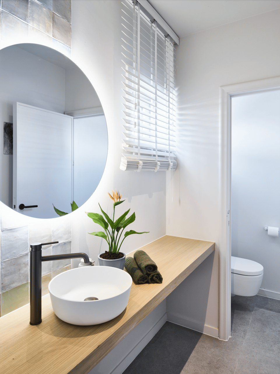 Moderne badkamer met ronde spiegel, wastafel op houten plank, plant en toilet.