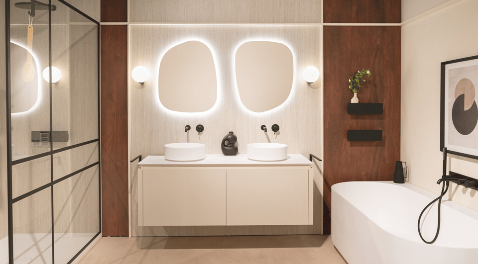 Salle de bains contemporaine de x²o salles de bains avec un meuble suspendu couleur crème, deux vasques rondes blanches mates et deux miroirs aux formes organiques avec éclairage LED indirect. Le mur arrière combine des panneaux clairs texturés et des panneaux en bois foncé. À gauche, une paroi de douche noire et à droite une baignoire îlot blanche avec robinetterie noire.