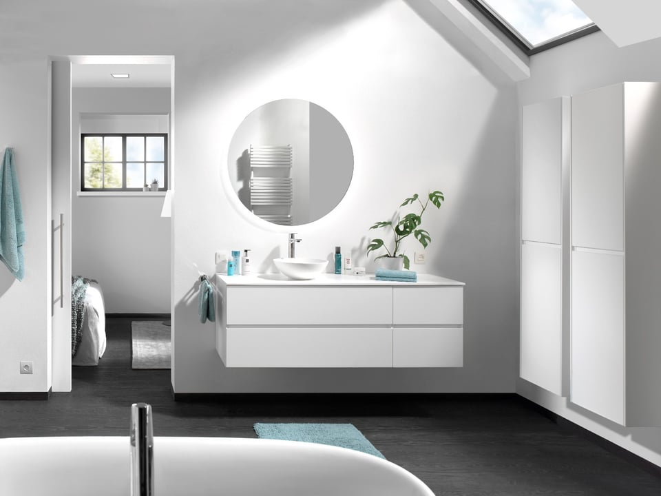 Salle de bains mansardée moderne avec meuble lavabo minimaliste, miroir rond, produits de soin et fenêtre de toit.