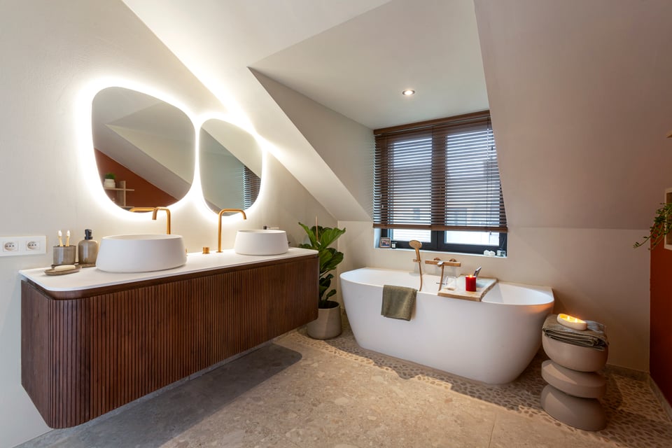 Een stijlvolle x²o badkamers ruimte met een vrijstaand bad, een houten badkamermeubel met twee opbouwkommen en ronde spiegels met geïntegreerde verlichting onder een schuin dak. Er staat een plant naast het bad en een stenen bijzettafel met kaarsjes.